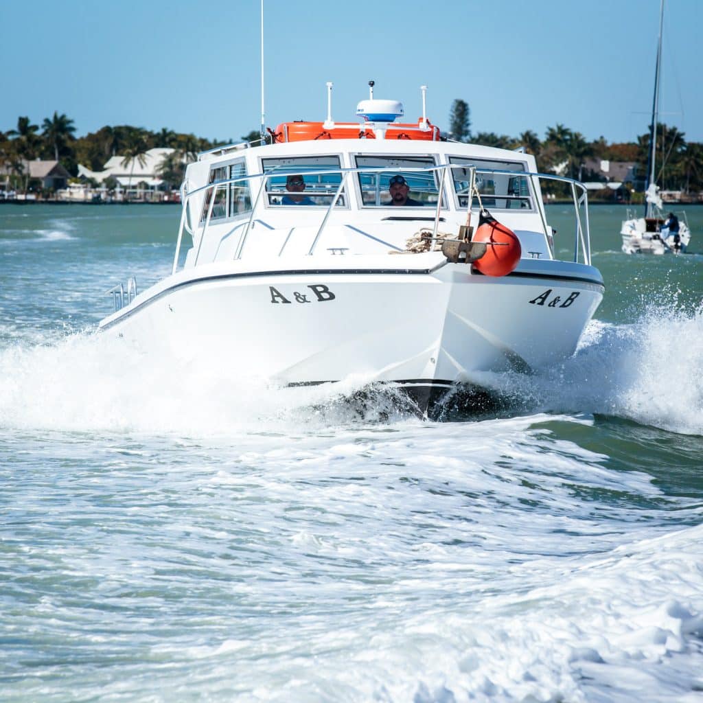 A white boat labeled A & B moves quickly through the water, creating waves on a sunny day. Two people are visible inside, likely enjoying a thrilling Naples FL Deep Sea Fishing adventure. The sky is clear, and houses and palm trees are in the background.