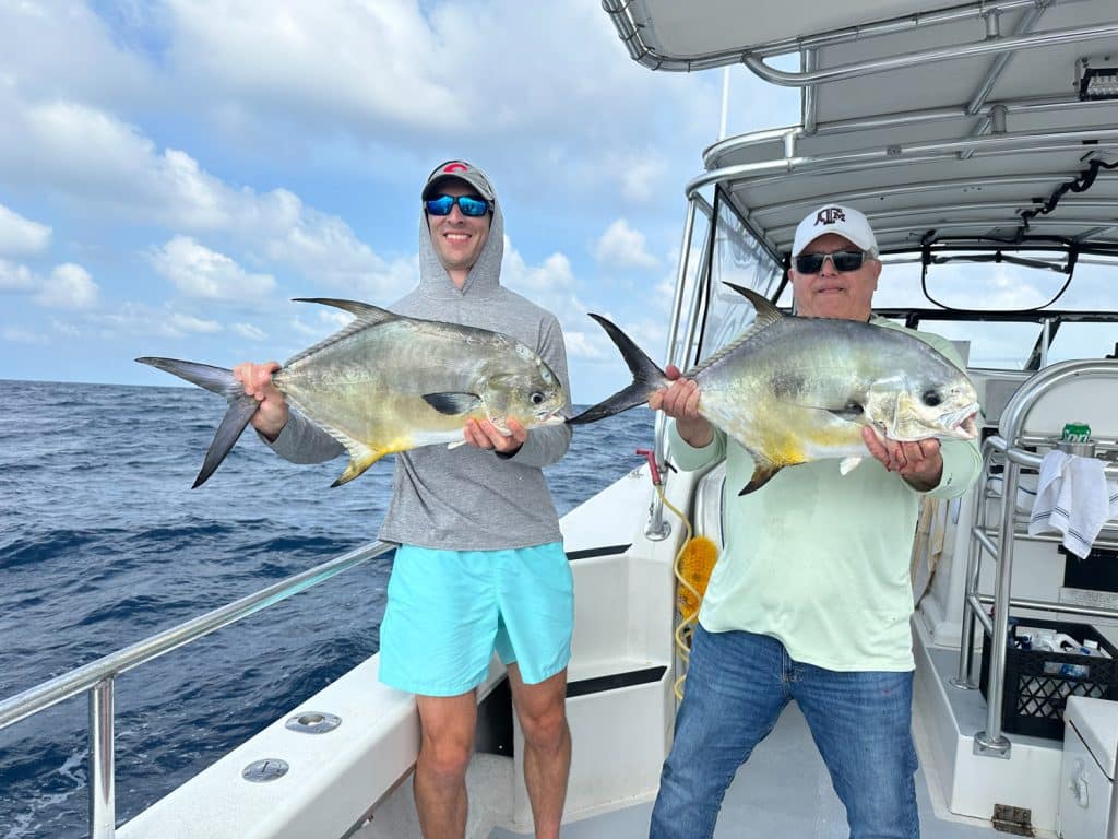 Two people on a boat holding large silver fish, smiling at the camera during their Naples Fishing Charter adventure. The ocean and a partly cloudy sky are in the background, capturing the thrill of Naples FL Deep Sea Fishing.