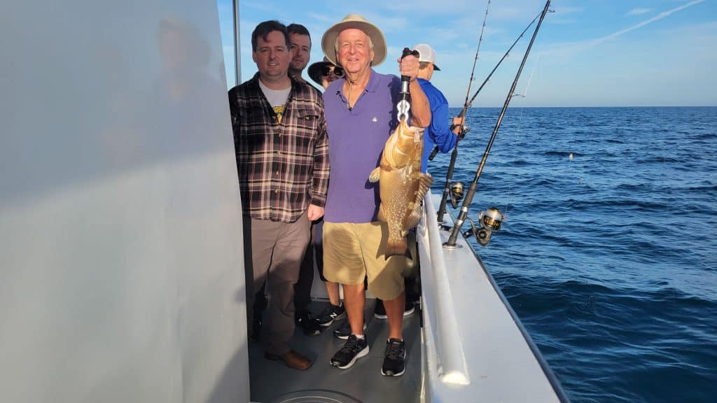 Three men stand on a boat holding fishing rods; one man in a hat and purple shirt proudly holds a large fish. The ocean and blue sky are visible in the background, capturing the excitement of a Naples FL Deep Sea Fishing adventure with a top-rated Naples Fishing Charter.