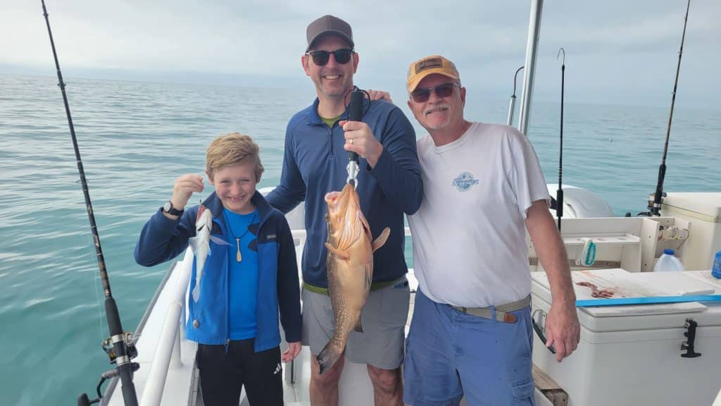 Three people on a boat hold up their catches—one boy with a small fish, two men with larger fish. They’re smiling with fishing gear and the ocean in the background under a cloudy sky, enjoying an unforgettable Naples FL Deep Sea Fishing experience.