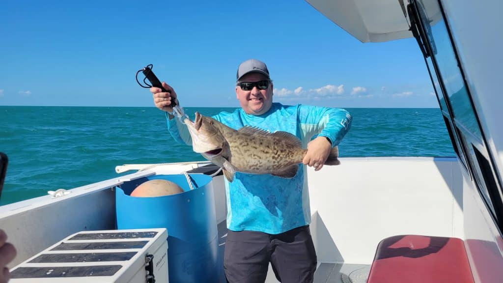 A man in sunglasses and a blue shirt holds a large fish on a boat during a Naples Fishing Charter, with the ocean and blue sky in the background.