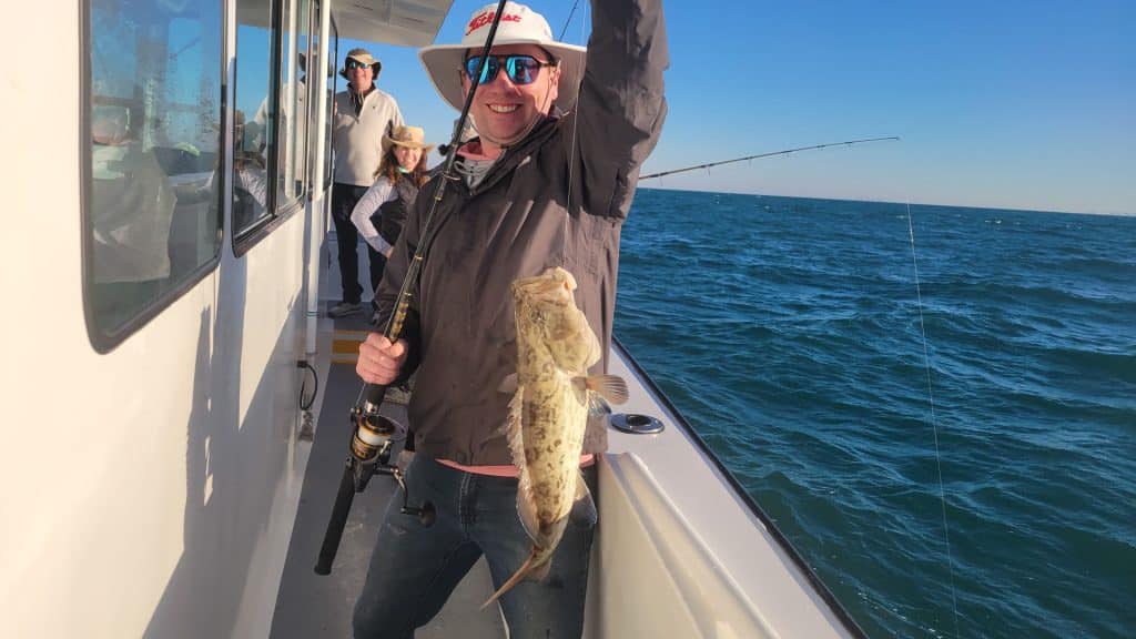 A man on a boat holds up a large fish he caught, smiling at the camera during an exciting Naples FL Deep Sea Fishing adventure. Two other people stand behind him, and the ocean is visible under a clear blue sky.