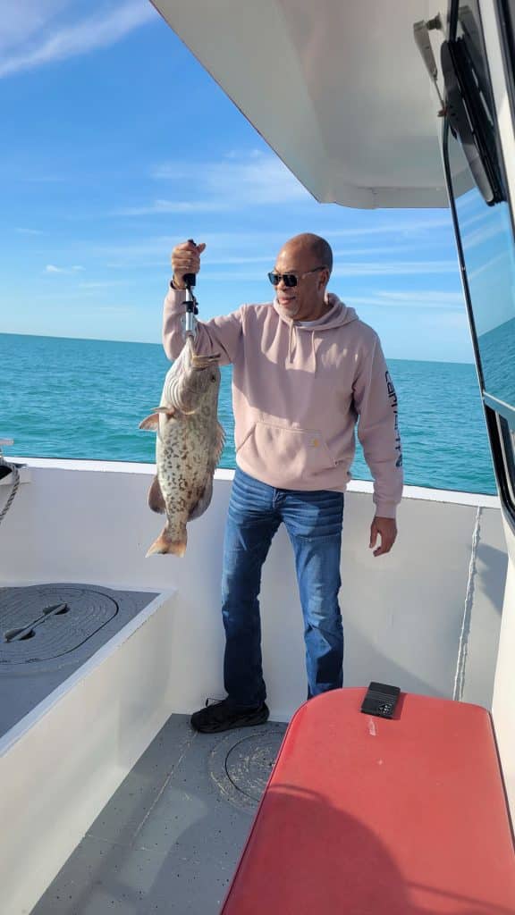 A man in sunglasses and a pink hoodie stands on a boat, smiling and holding a large fish with a tool during his Naples FL Deep Sea Fishing adventure. The ocean and blue sky stretch out in the background, perfectly capturing the excitement of a Naples Fishing Charter.