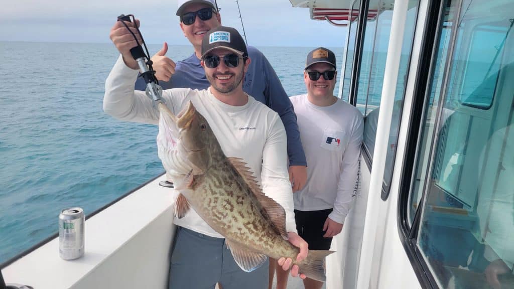 Three men on a boat smile at the camera as one holds up a large fish, showcasing their successful Naples FL Deep Sea Fishing adventure. Another man gives a thumbs up, while the sparkling ocean stretches out in the background, capturing the excitement of their Naples Fishing Charter experience.