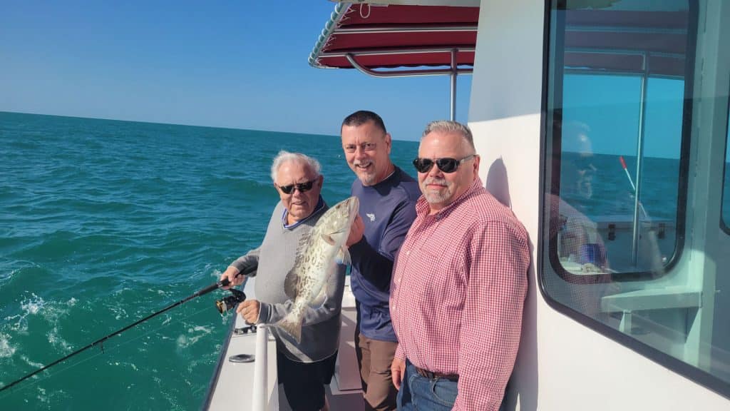 Three men stand on a boat in the ocean, enjoying an unforgettable Naples FL Deep Sea Fishing experience. One holds a fishing rod with a large fish, while all three smile at the camera under a sunny sky.
