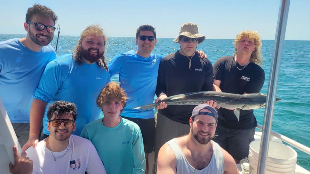Eight men are on a boat at sea, smiling at the camera during an unforgettable Naples FL Deep Sea Fishing adventure. One man is proudly holding a large fish, while the sunny weather, clear skies, and calm waters provide the perfect backdrop for this exciting Naples Fishing Charter experience.