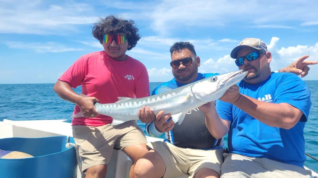 Three people on a boat, enjoying a Naples FL Deep Sea Fishing adventure, hold a large silver fish together and smile at the camera. The sky is bright blue with a few clouds, and the ocean stretches out in the background.