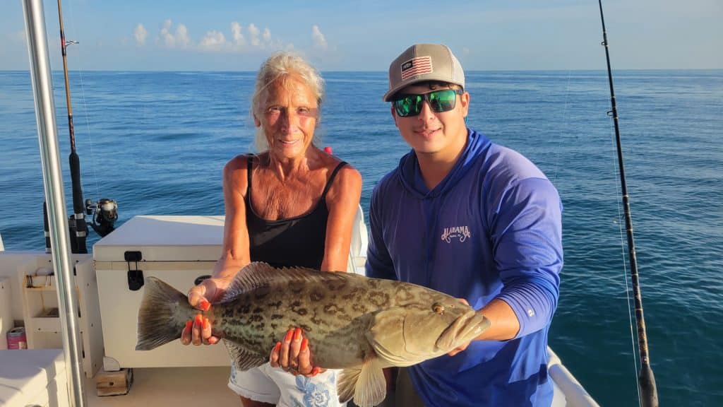 Two people on a boat hold a large fish together, smiling at the camera after an exciting Naples FL Deep Sea Fishing adventure. The ocean and clear sky are in the background, with fishing rods visible on the right side, capturing the thrill of a memorable Naples Fishing Charter.