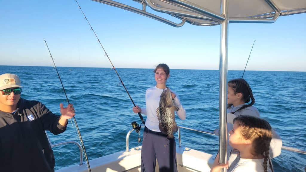 Four people are on a boat at sea with a backdrop of clear, blue skies. One person in the center smiles and proudly holds a large fish, while others stand nearby, enjoying the moment together. Fishing rods are visible on this exciting Naples FL Deep Sea Fishing adventure, making it a memorable Naples Fishing Charter for everyone onboard.