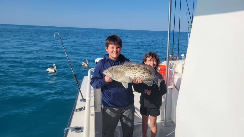 Two kids on a boat hold a large fish together, smiling at the camera during their Naples FL Deep Sea Fishing adventure. Fishing rods are nearby, and pelicans float on the blue sea under a clear sky, capturing the excitement of a Naples Fishing Charter.