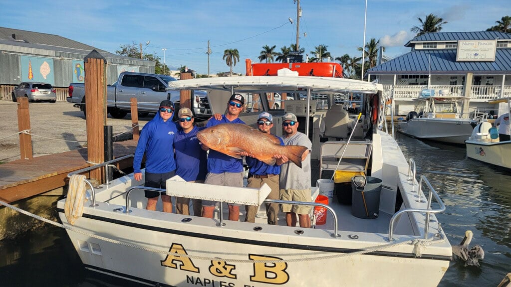 Five people on a boat at a marina proudly hold a large fish together. The boat, with A & B Naples written on the back, hints at an exciting Naples Fishing Charter adventure. It’s a sunny day with buildings, palm trees, and other boats in the background—a perfect snapshot of Naples FL Deep Sea Fishing fun.