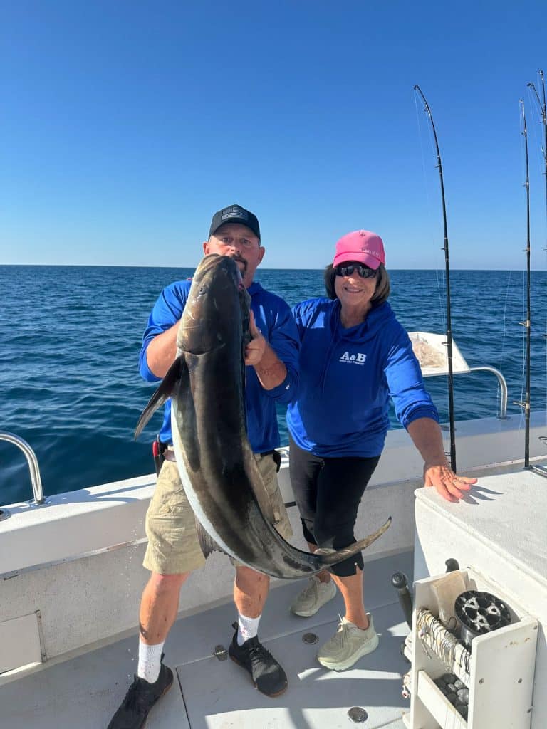 Two people smile on a boat under a clear sky, holding a large fish together while enjoying their Naples Fishing Charter. Fishing rods are visible nearby, and the calm ocean in the background sets the perfect scene for an unforgettable Naples FL Deep Sea Fishing adventure.