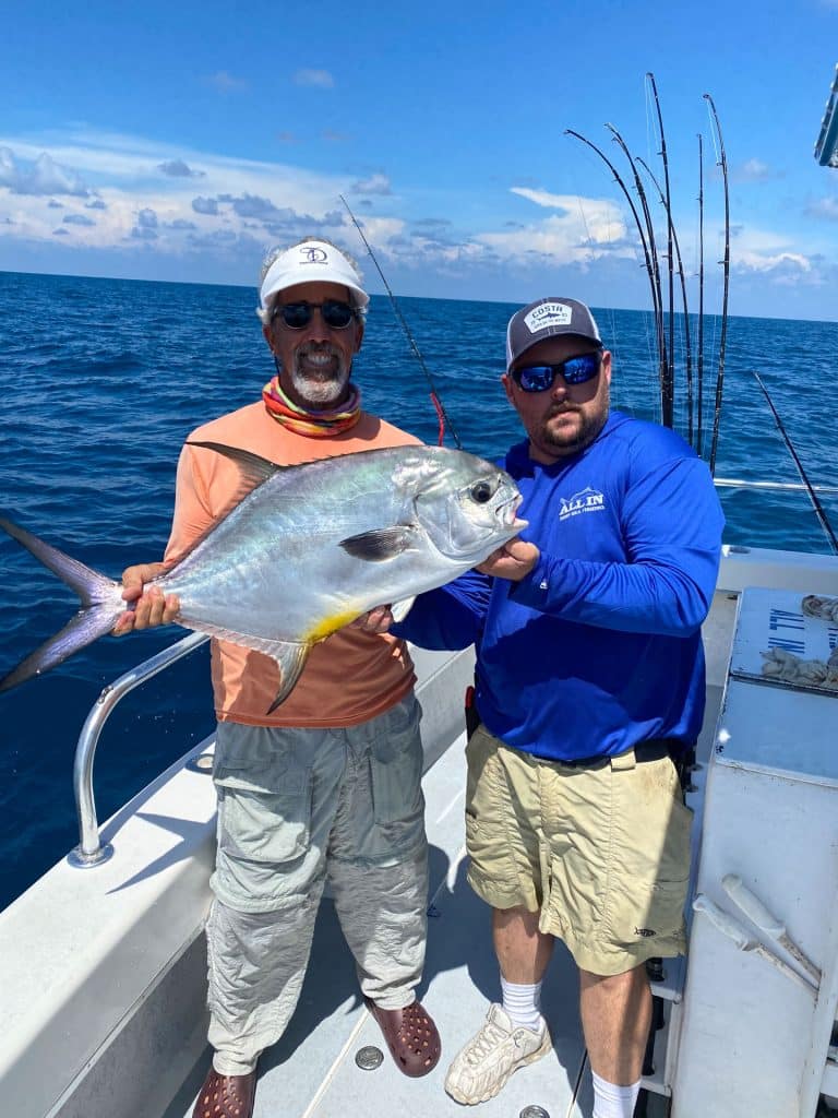 Two men on a boat at sea proudly hold a large silver fish during their Naples FL Deep Sea Fishing adventure. Fishing rods are visible in the background under a partly cloudy sky.