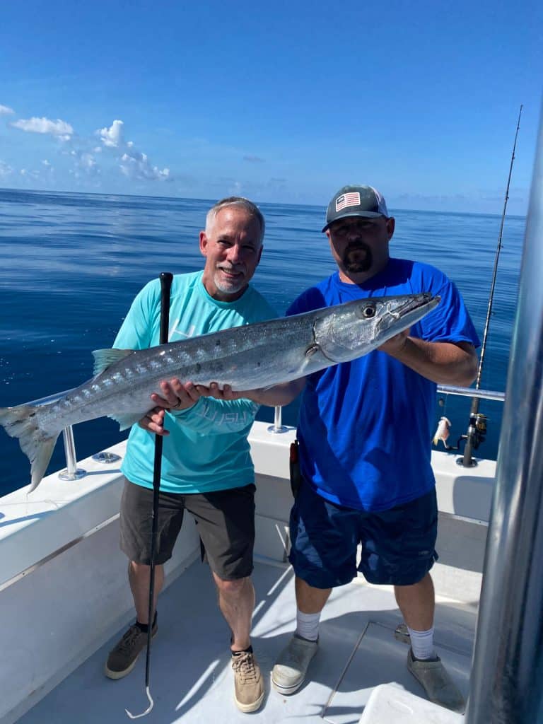 Two men on a boat hold a large fish together, smiling at the camera during their Naples FL Deep Sea Fishing adventure. The ocean is calm and blue in the background, with a clear sky overhead. One man wears a teal shirt, the other wears a blue shirt and cap—both enjoying an unforgettable day with a Naples Fishing Charter.