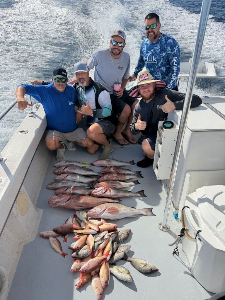 Four men on a fishing boat pose with a large catch of fish laid out on the deck, proudly showing off their success from an exciting Naples FL Deep Sea Fishing adventure. They smile and give thumbs up with the ocean wake visible behind them on a sunny day, capturing the thrill of a memorable Naples Fishing Charter.