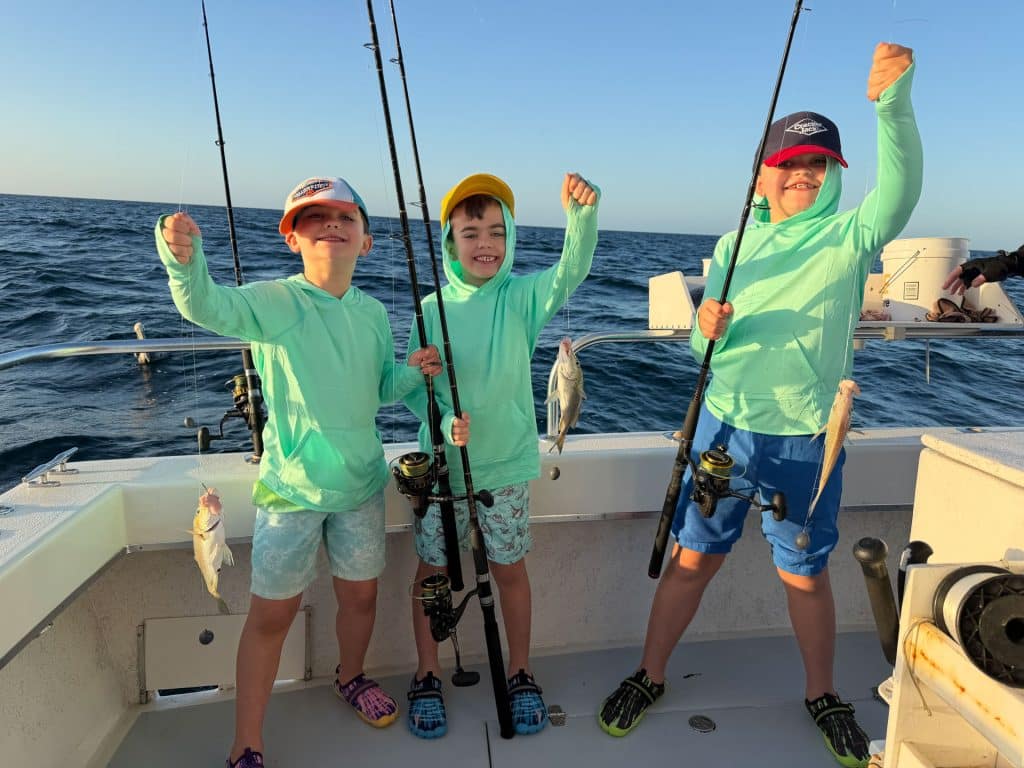 Three children on a boat, wearing light green hoodies and caps, smile and hold fishing poles with small fish they’ve caught during an exciting Naples FL Deep Sea Fishing adventure. The ocean and clear sky are visible in the background.