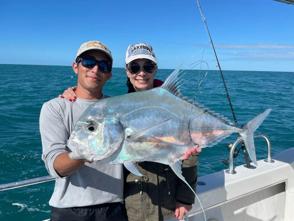 Two people on a boat holding a large, shiny fish with long fins during their Naples FL Deep Sea Fishing adventure. The ocean and blue sky are in the background, while both are wearing hats and sunglasses.