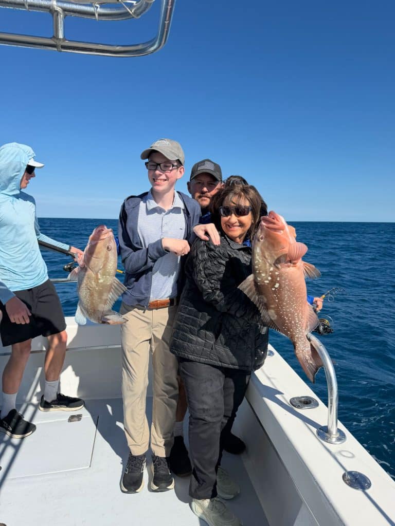 Four people on a boat, two holding large fish and smiling, enjoying a memorable Naples FL Deep Sea Fishing adventure. The ocean and blue sky are in the background, capturing the excitement of their Naples Fishing Charter experience.