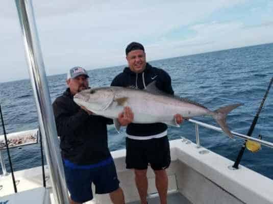 Two men on a boat holding a large fish together, with the ocean and cloudy sky in the background—capturing an exciting moment from a Naples FL Deep Sea Fishing adventure.