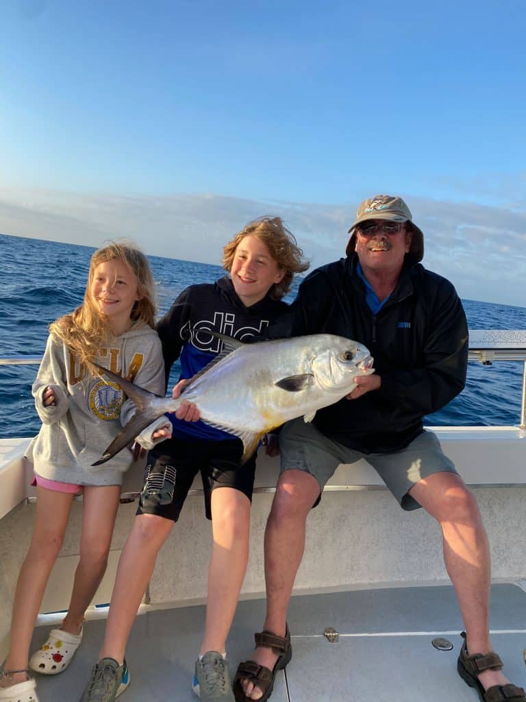 Three people sit on a boat, smiling and holding a large fish during their Naples FL Deep Sea Fishing adventure. The ocean is visible in the background under a blue sky with clouds.