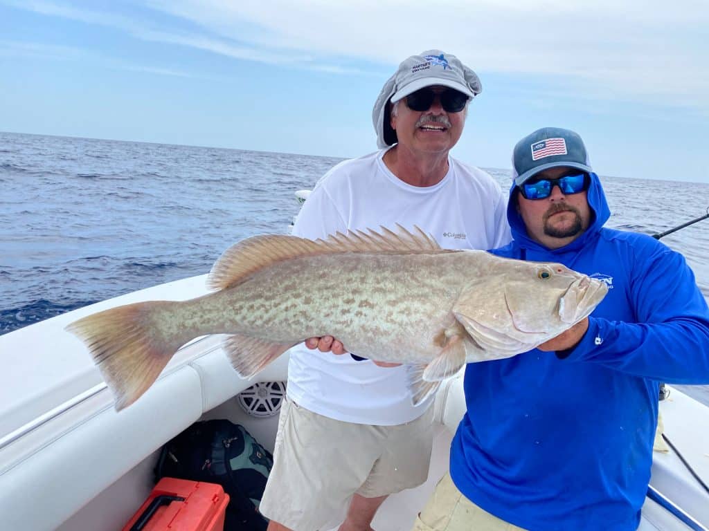 Two men on a boat hold a large, light-colored fish together during their Naples Fishing Charter adventure. The man on the left wears a white shirt and sunglasses; the man on the right wears a blue hoodie and hat. The ocean and sky provide a scenic backdrop, capturing an unforgettable moment of Naples FL Deep Sea Fishing.