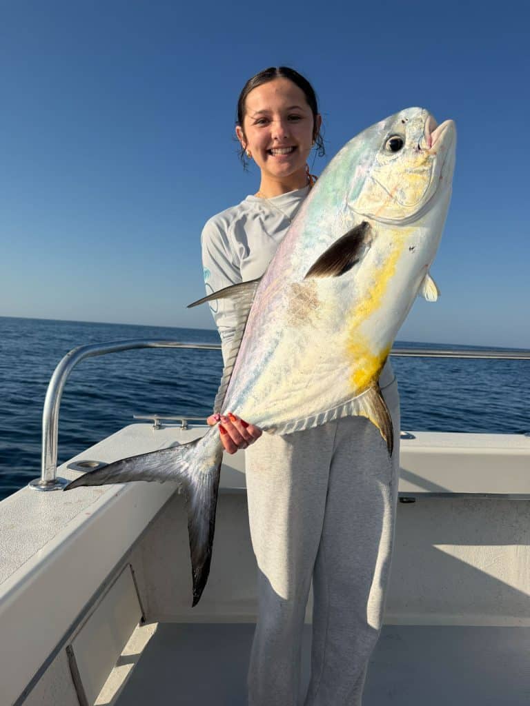 A smiling person on a Naples Fishing Charter holds a large, silvery fish with yellow markings against a clear blue sky and calm ocean background.
