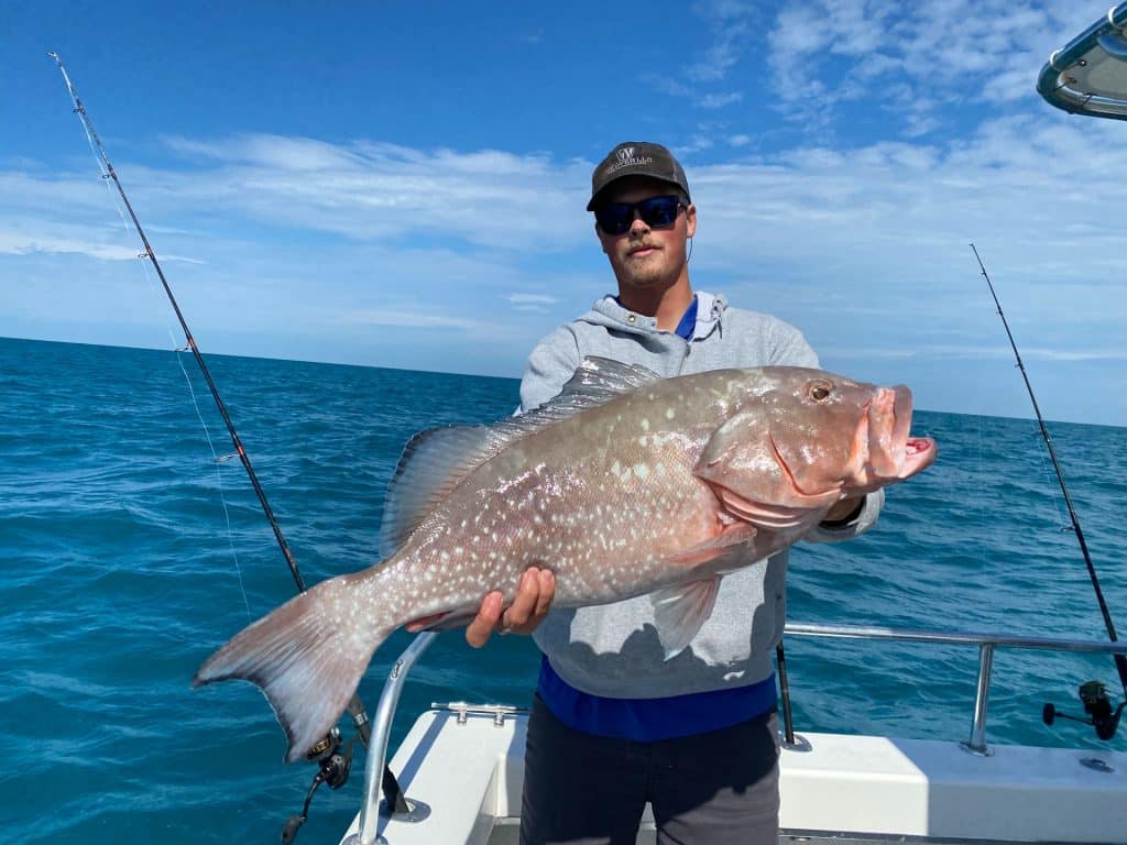 A person on a boat holding a large grouper fish, with fishing rods nearby and the ocean and blue sky in the background, showcases the excitement of a Naples FL Deep Sea Fishing adventure.