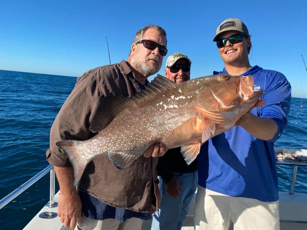 Three men on a boat hold a large fish together, smiling at the camera. The ocean and blue sky are in the background, with fishing rods visible behind them—capturing the excitement of a successful day with a Naples Fishing Charter.