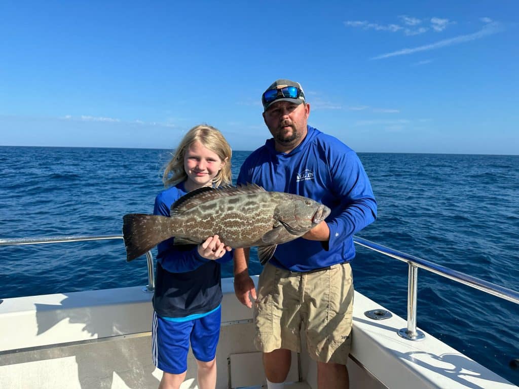 A child and an adult on a Naples Fishing Charter boat hold a large fish together, with the ocean and blue sky in the background. Both are smiling, wearing blue tops, and standing near the boat’s railing.