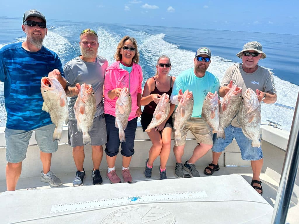 Six people on a boat holding up large fish they caught with Naples FL Deep Sea Fishing, smiling at the camera with the ocean and wake visible behind them under a sunny, blue sky.
