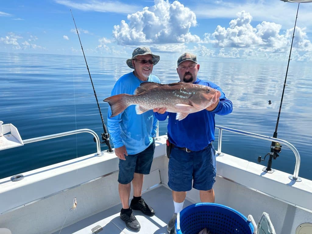 Two men on a boat holding a large fish together, with fishing rods in the background and calm water under a blue sky with clouds, capture the excitement of a Naples FL Deep Sea Fishing adventure.