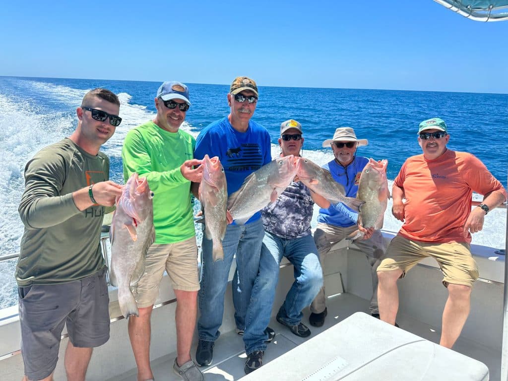 Six men on a boat hold up large fish they caught with a Naples Fishing Charter, smiling on a sunny day with blue sky and ocean in the background.