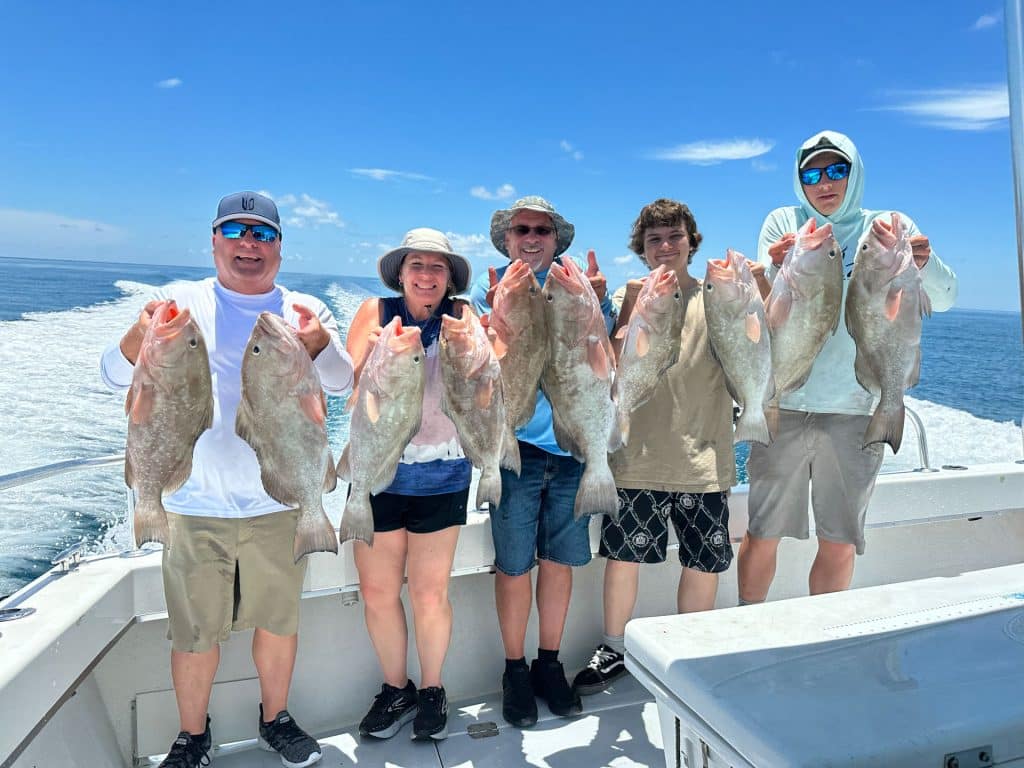 Five people on a boat hold large fish, smiling at the camera. Blue sky and ocean are in the background, with a trail of water behind the moving boat—a perfect day out with a Naples Fishing Charter.