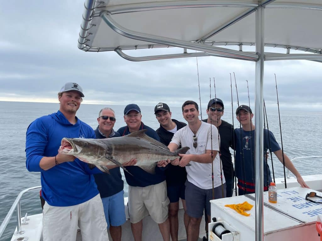 Six men on a boat smile and hold a large fish together, with fishing rods in the background and the ocean visible behind them under a cloudy sky—capturing an unforgettable moment during their Naples Fishing Charter adventure.