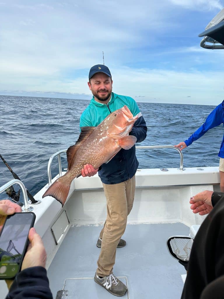 A man on a boat holds a large fish, smiling at the camera during an exciting Naples FL Deep Sea Fishing adventure. The boat is on the ocean, with blue sky and clouds in the background. Others from the Naples Fishing Charter are partially visible on the boat.