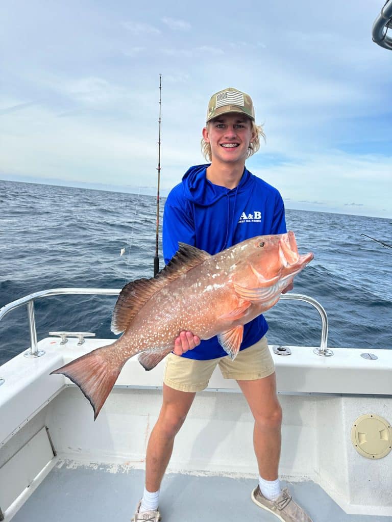 A young person wearing a blue hoodie and cap stands on a boat, smiling and holding a large fish with the ocean and a fishing rod visible in the background—capturing the excitement of a Naples FL Deep Sea Fishing adventure.