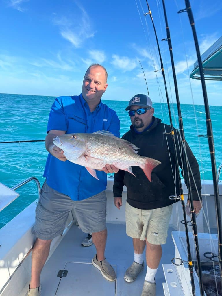 Two men on a boat at sea, one holding a large fish and smiling after a successful Naples FL Deep Sea Fishing trip. The other stands beside him wearing sunglasses and a cap. Several fishing rods are visible, and the water is bright blue under a clear sky.