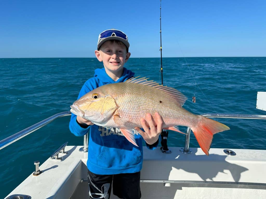 A smiling child in a blue hoodie holds a large fish on a boat during an exciting Naples FL Deep Sea Fishing adventure, with blue ocean water and a fishing pole in the background under a clear sky.