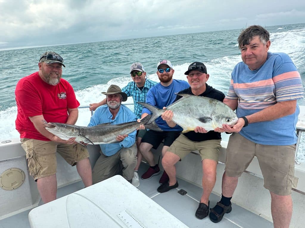 Six men on a boat pose with two large fish they caught during their Naples Fishing Charter, smiling at the camera with the vast ocean and a cloudy sky in the background.
