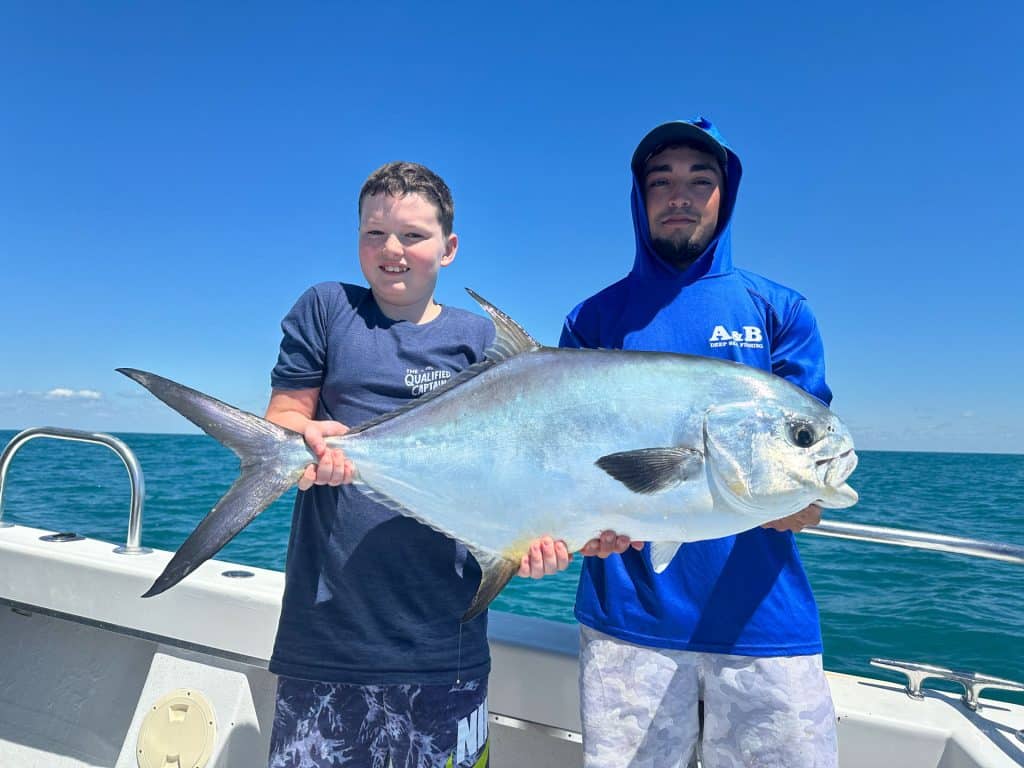 Two people on a boat hold a large silver fish, beaming with pride after an exhilarating Naples Fishing Charter. The ocean and clear blue sky in the background highlight their unforgettable Naples FL Deep Sea Fishing adventure.