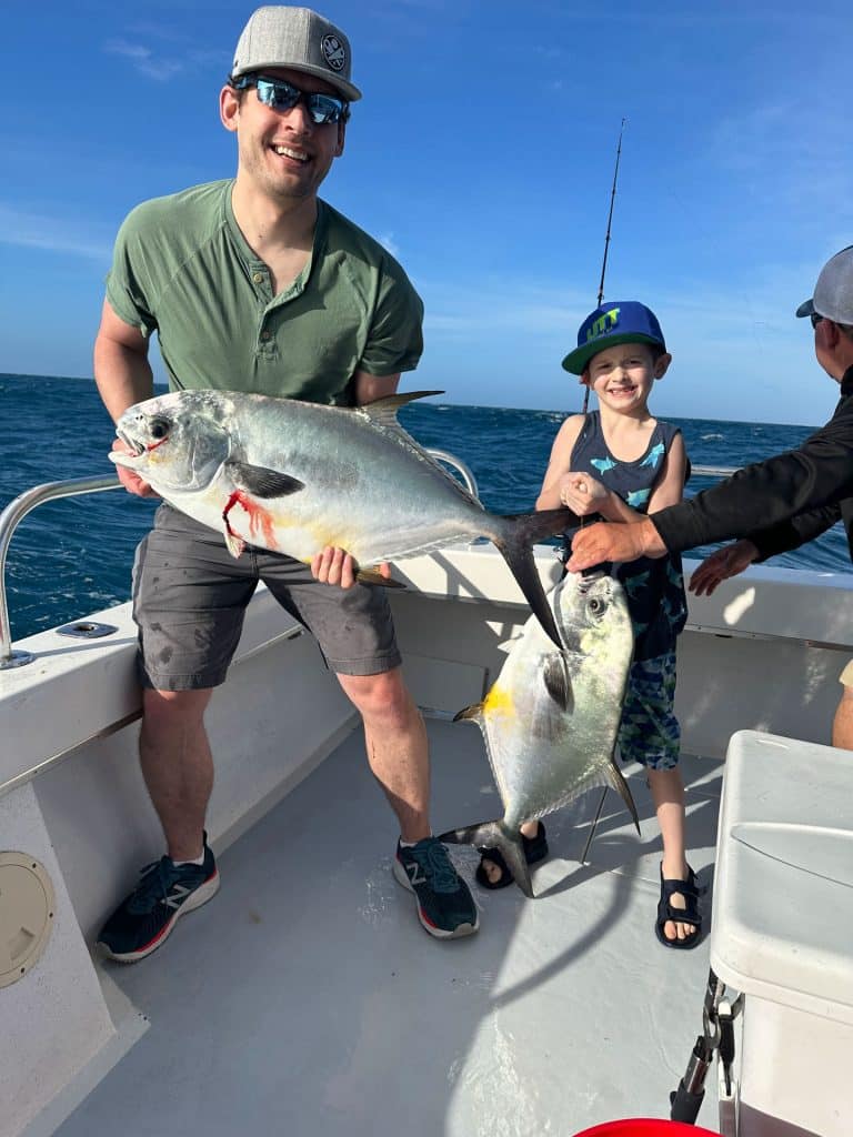 A man and a young boy on a boat smile while each holds a large fish, enjoying their Naples FL Deep Sea Fishing adventure. The ocean and blue sky are in the background, with fishing rods from their Naples Fishing Charter visible.
