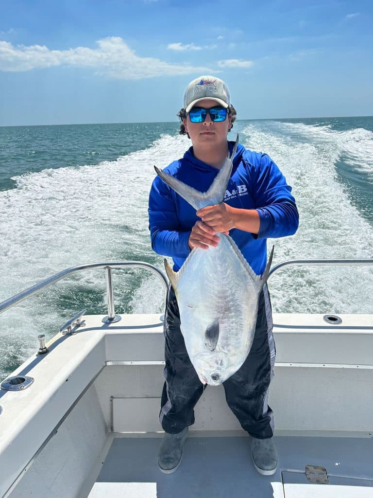 A person in a blue shirt and cap holds a large silver fish on a boat, with the ocean and wake visible behind them under a sunny sky—a perfect moment captured during a Naples Fishing Charter adventure.