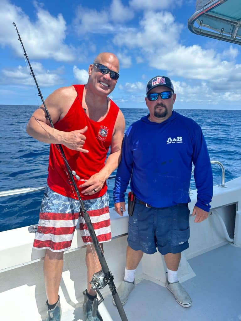 Two men pose on a boat under a blue sky during their Naples FL Deep Sea Fishing adventure. One holds a fishing rod and wears a red tank top and striped shorts; the other, dressed in a blue long-sleeve shirt and sunglasses, stands beside him. The expansive ocean forms the backdrop of this unforgettable Naples Fishing Charter experience.