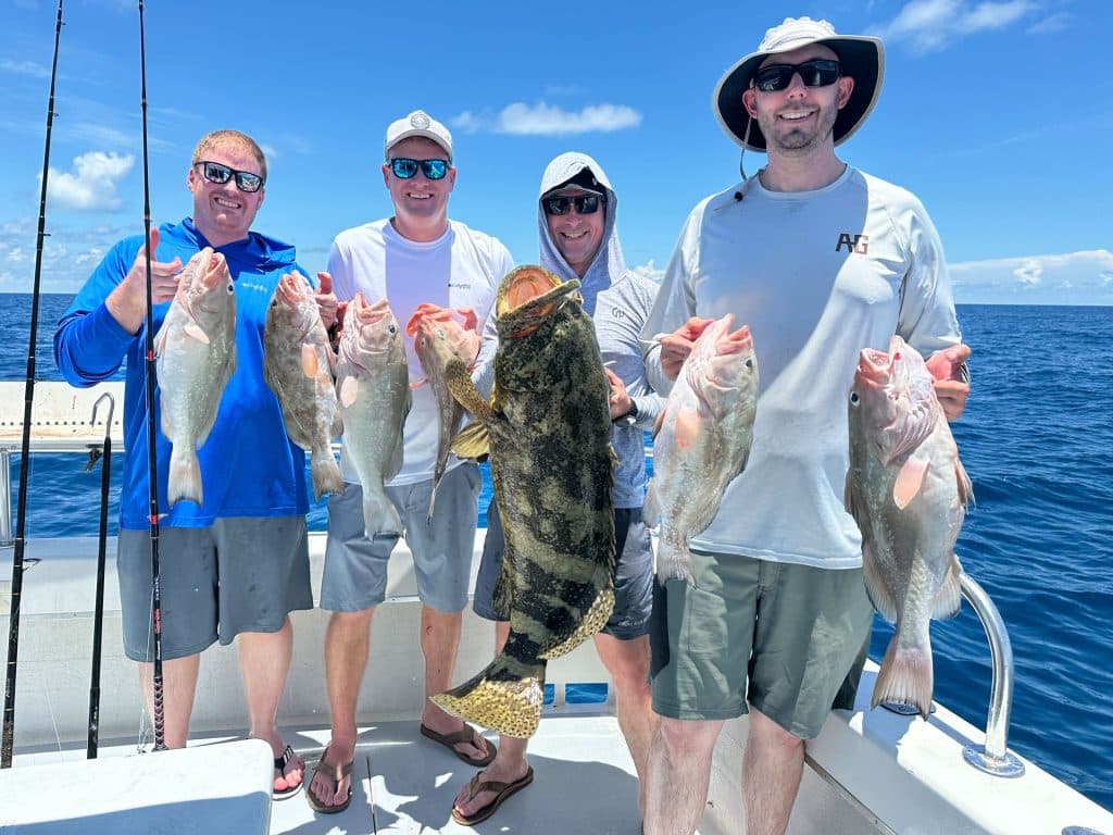 Four men on a boat holding freshly caught fish, including a large grouper, smiling under a sunny blue sky with ocean in the background—an unforgettable day with Naples Fishing Charter.
