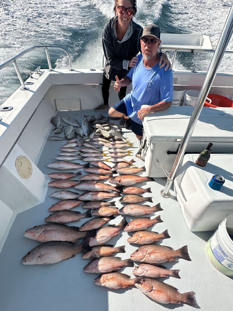 Two people on a boat pose with their catch: about twenty-five fish laid out in neat rows on the boat’s deck, the ocean visible behind them—a successful outing with a Naples FL Deep Sea Fishing charter.