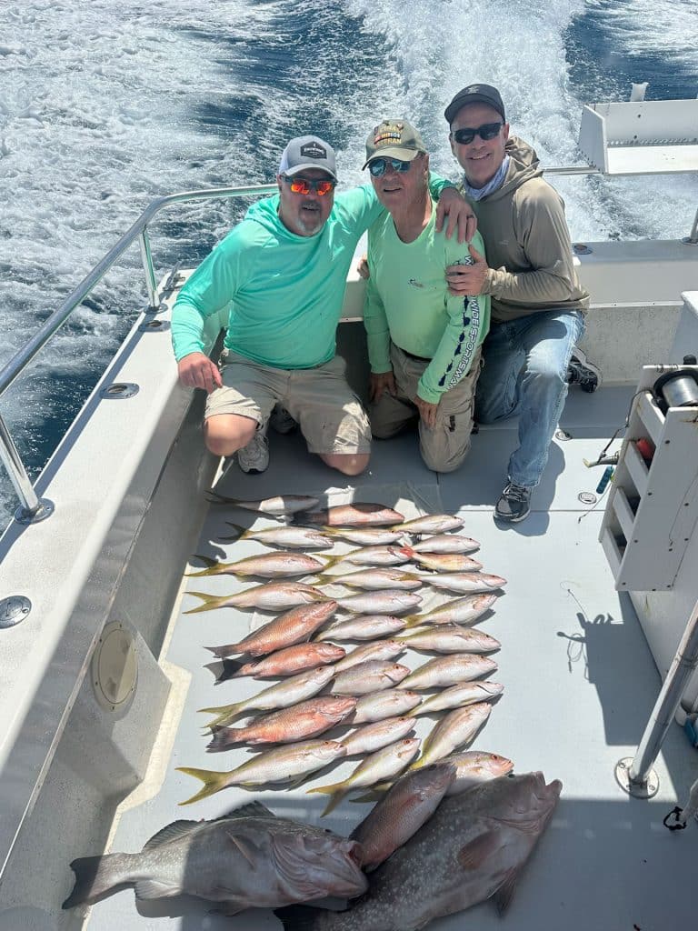 Three men pose on a Naples Fishing Charter boat with a large catch of fish neatly arranged on the deck. The ocean waves trail behind the boat, and all three are smiling at the camera under bright sunlight.