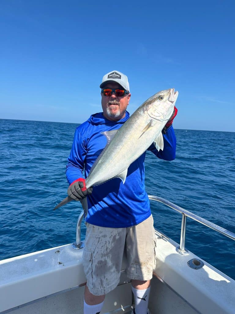A man wearing sunglasses, a blue shirt, and a cap holds a large fish on a boat with the ocean and clear blue sky in the background, capturing the thrill of a Naples Fishing Charter adventure.
