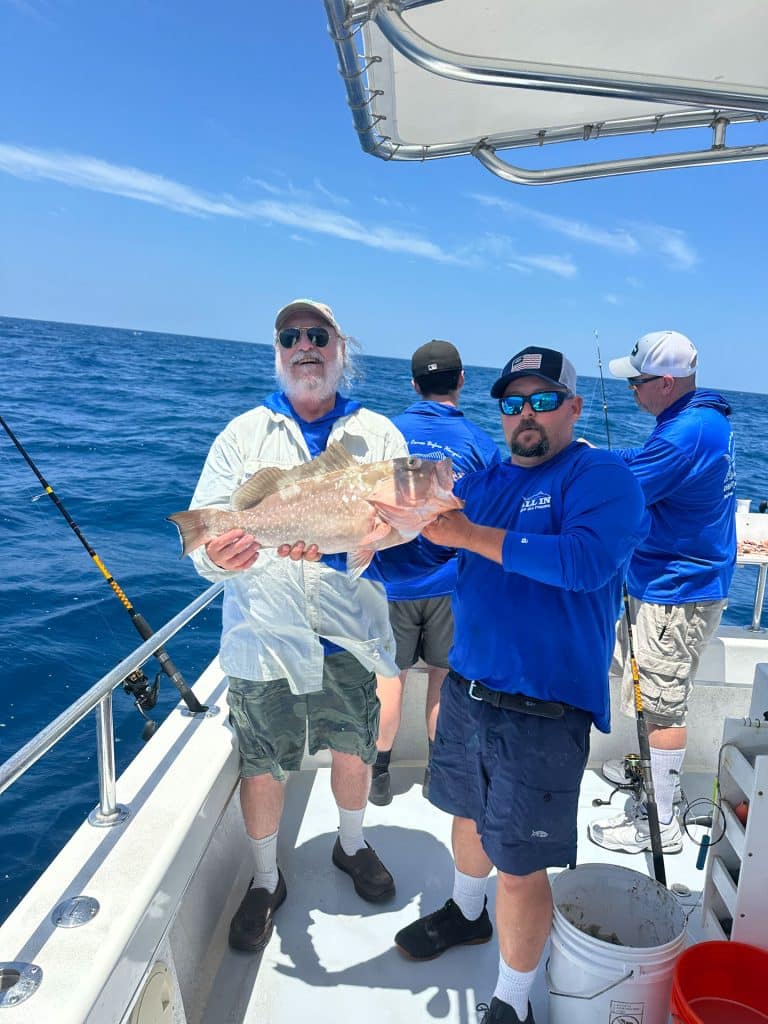 Two men on a boat proudly hold a large fish, while two others face the ocean, fishing. The sky is clear and the water is calm, suggesting a pleasant day at sea—perfect for an unforgettable Naples FL Deep Sea Fishing adventure with an experienced Naples Fishing Charter.