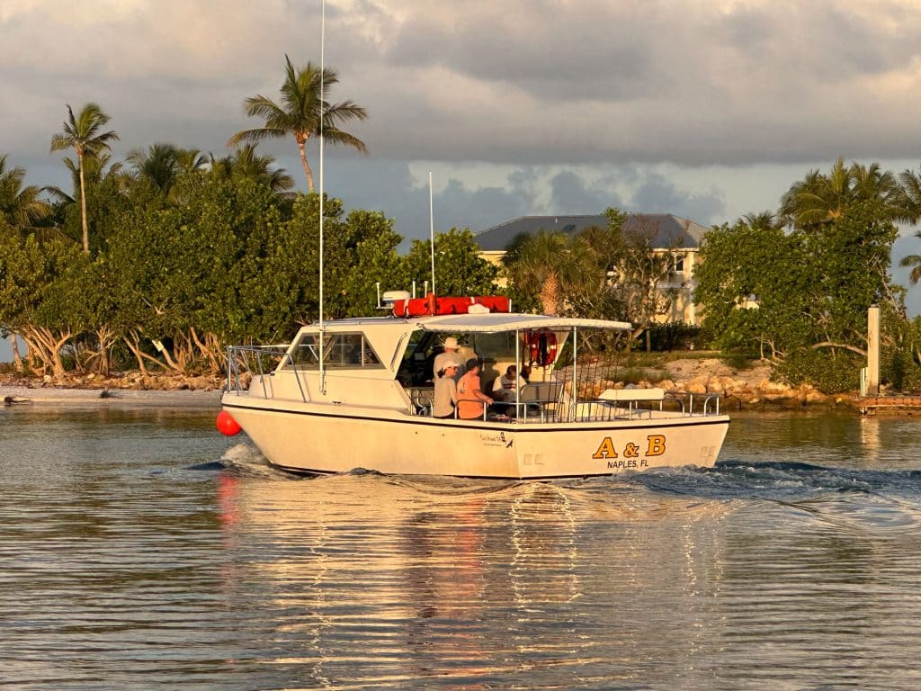 A white boat labeled A & B Naples FL moves through calm water near trees and a house, with two people visible onboard under a partly cloudy sky, capturing the essence of a Naples Fishing Charter adventure.