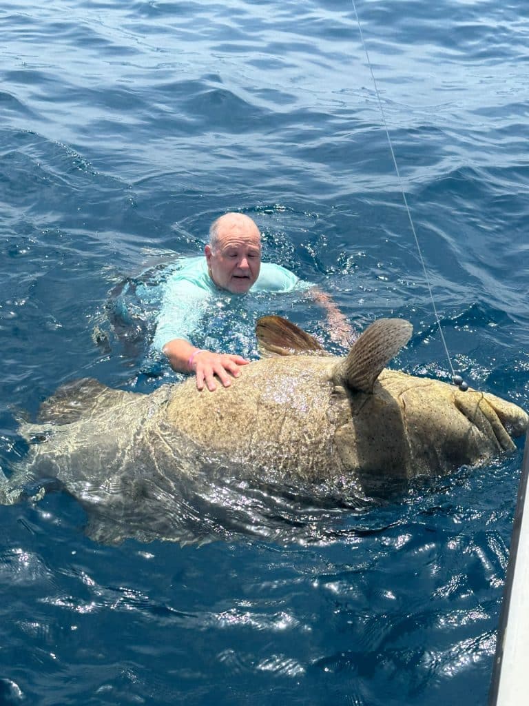 An older man in a light blue shirt is in the ocean, holding onto a large fish beside a boat—an unforgettable moment on a Naples FL Deep Sea Fishing adventure. The water is deep blue, and both the man and fish are partially above the water’s surface.